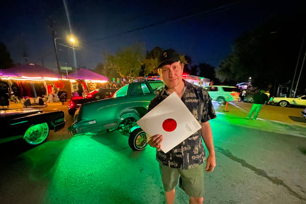 A man holds up a record in front of a green-lit lowrider. It's dark and he's standing in a parking lot.